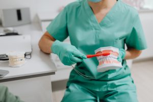 a dental professional wearing green scrubs showing a patient how to properly brush their teeth on a dental model.